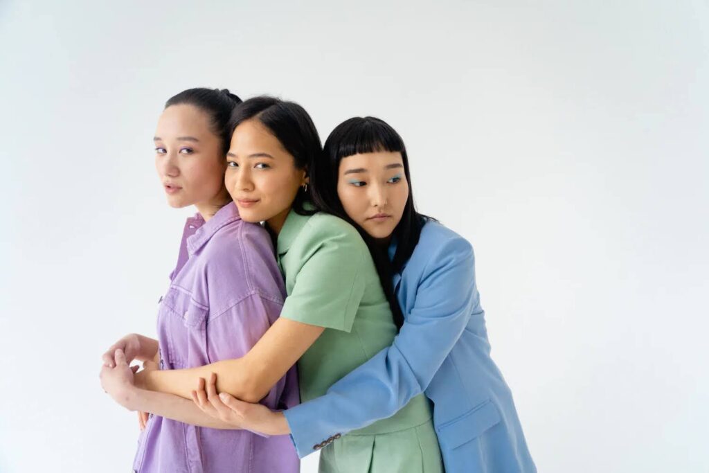 Three young women of Asian descent are posing together. The woman in the middle is wearing a purple shirt and has her arms wrapped around the other two women.