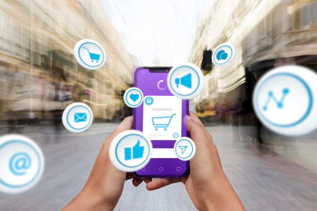 A person is holding a smartphone in their hand. The phone has a purple screen with a shopping cart icon on it. There are also several other icons surrounding the phone, such as a heart, a thumbs-up, and a chat bubble.