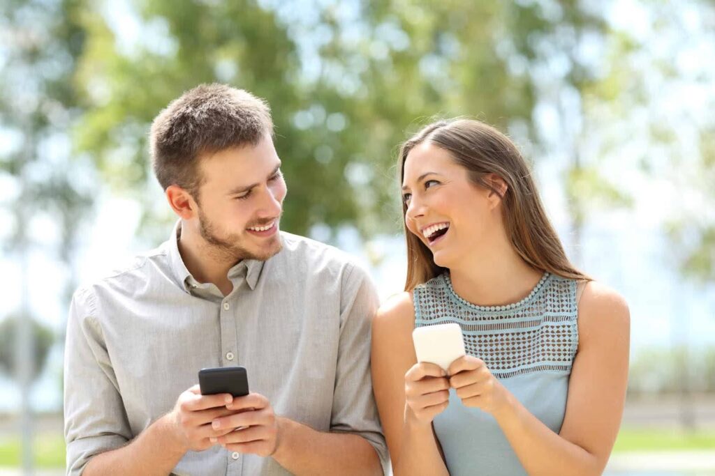 The picture shows a young man and woman standing next to each other, both looking at their phones and smiling.