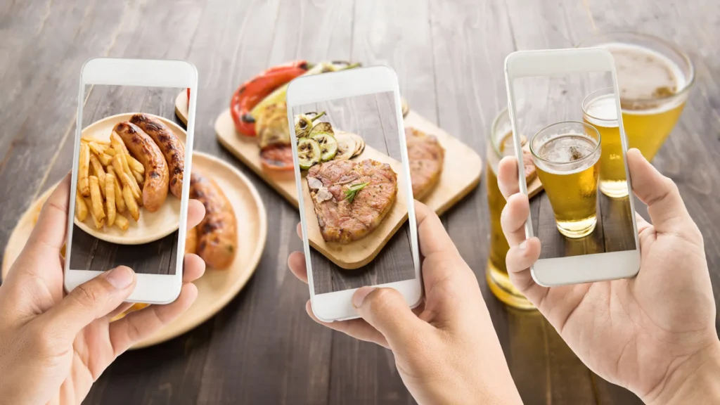 Three people are photographing meals and drinks on their smartphones. Meals include sausages with fries, grilled vegetables, meat patties, and beer on a table with a wooden surface.