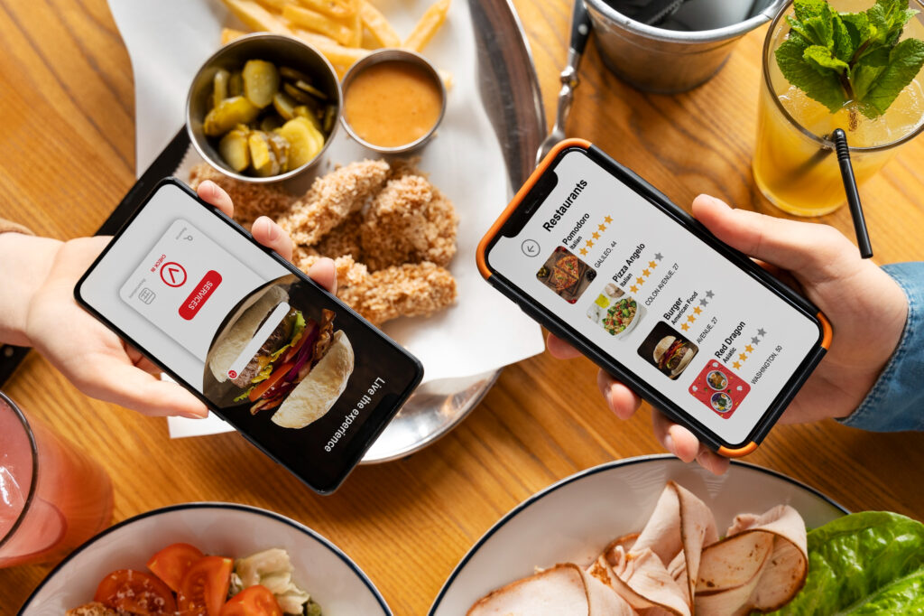 Two people holding smartphones displaying food delivery apps above a table with various dishes, including fried chicken, pickles, sauce, and drinks.