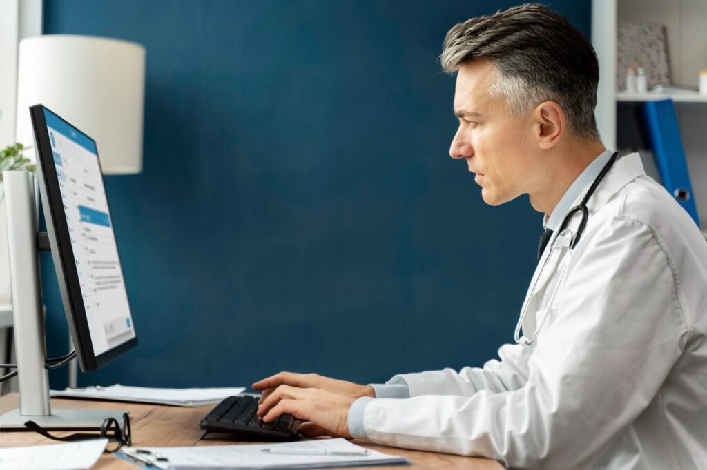 A doctor in a white coat and stethoscope around his neck types on a computer keyboard while looking at the monitor.